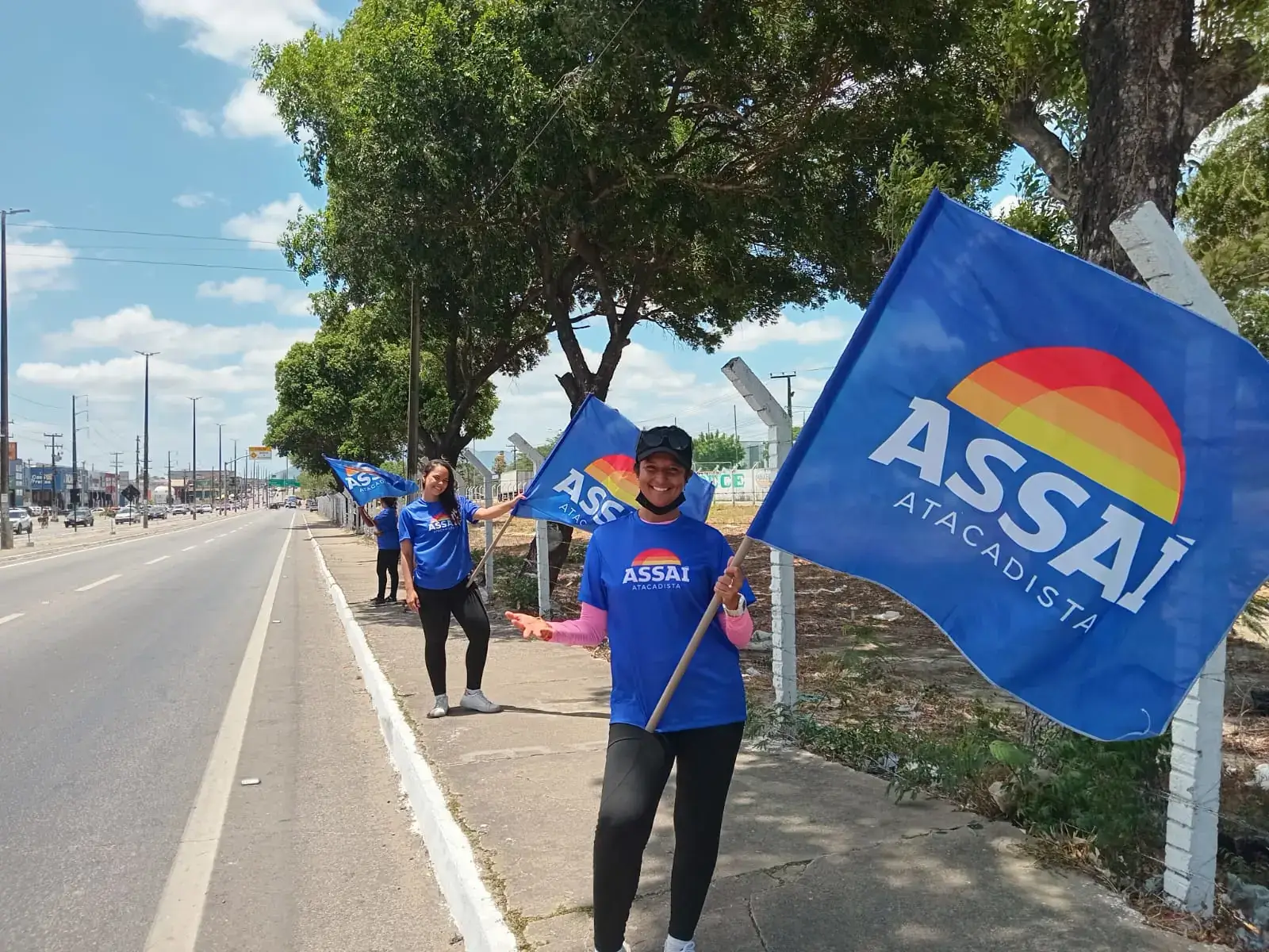 Ação de marketing de rua, conhecida como 'bandeiraço', do Assaí Atacadista. Promotoras de evento uniformizadas agitam bandeiras com o logo da marca na calçada de uma avenida movimentada para divulgar o supermercado.